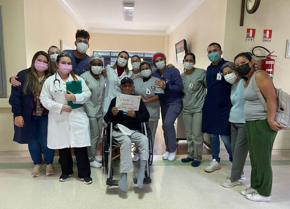 Carlos Massatoshi Higa posando com a equipe do hospital após receber alta. ele está na cadeira de rodas e segura um papel escrito "venci a covid-19"
