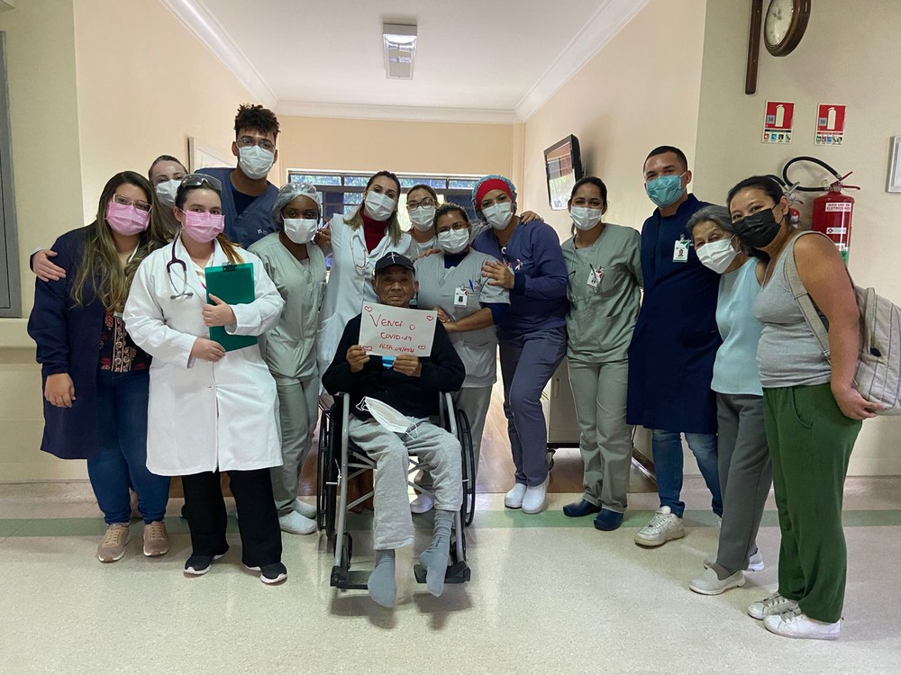 Carlos Massatoshi Higa posando com a equipe do hospital após receber alta. ele está na cadeira de rodas e segura um papel escrito "venci a covid-19"
