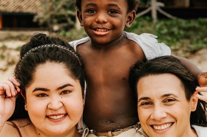 carolina e thais sorrindo para a foto com seu filho, daniel, sentado em seus ombros de camisa aberta