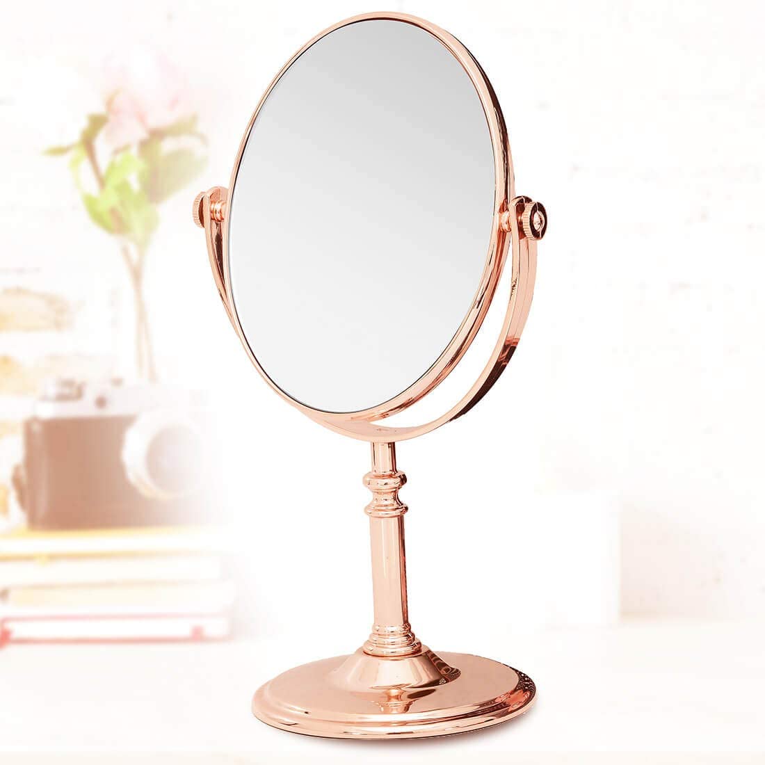 Em uma mesa, com algumas decora&ccedil;&otilde;es ao fundo, como plantas e livros, h&aacute; um espelho de mesa de maquiagem redondo. Ele gira para cima e para baixo. A estrutura &eacute; um rosa misturado com dourado