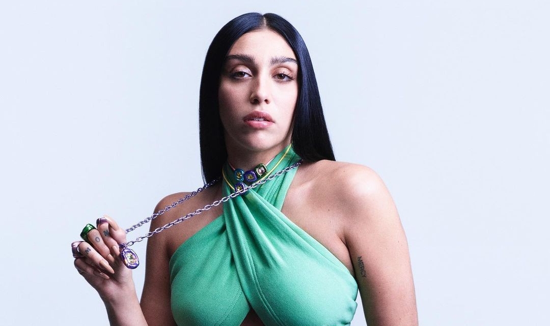 Imagens de Lourdes com top verde e calça jeans. Ela segura um colar que usa