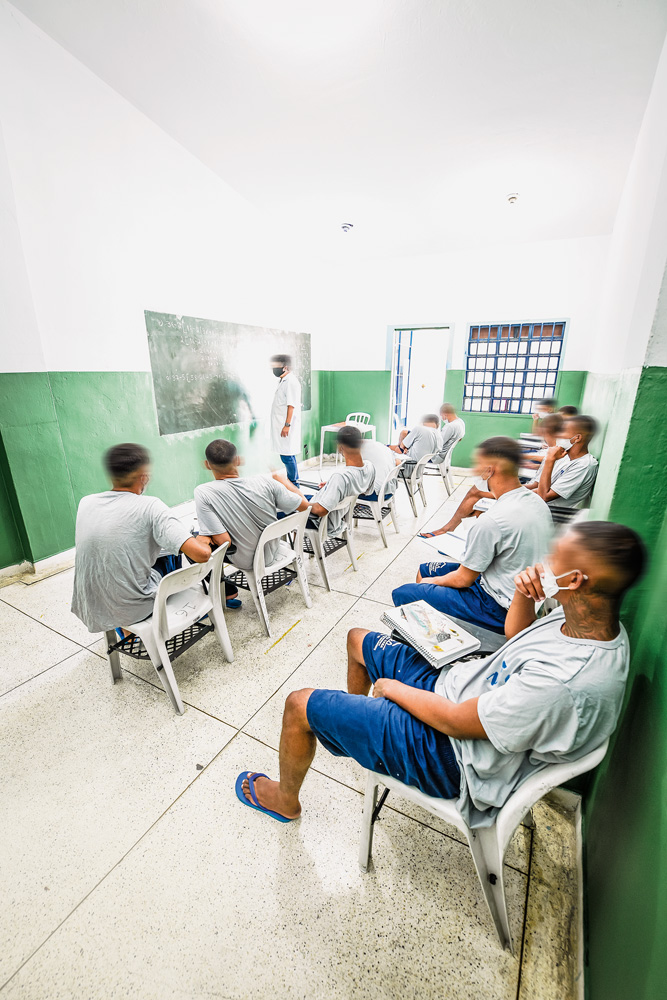 A imagem mostra alunos sentados em cadeiras de plásticos, vendo um professor na frente da sala, escrevendo em uma lousa.