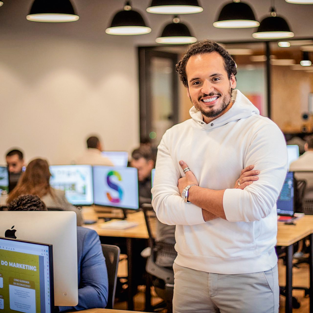 jovens-corretores-de-luxo-sp-Bruno-Sindona Bruno Sindona sorrindo de braços cruzados com computadores e pessoas trabalhando ao fundo