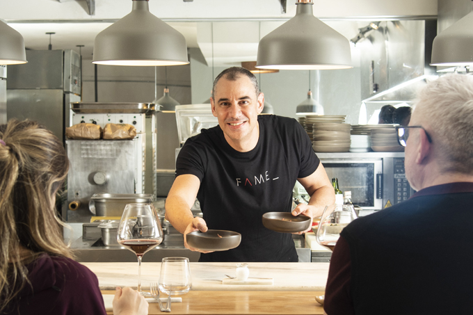 O chef Marco Renzetti atrás do bancada da cozinha do Fame entregando pratos para casal sentado à sua frente.