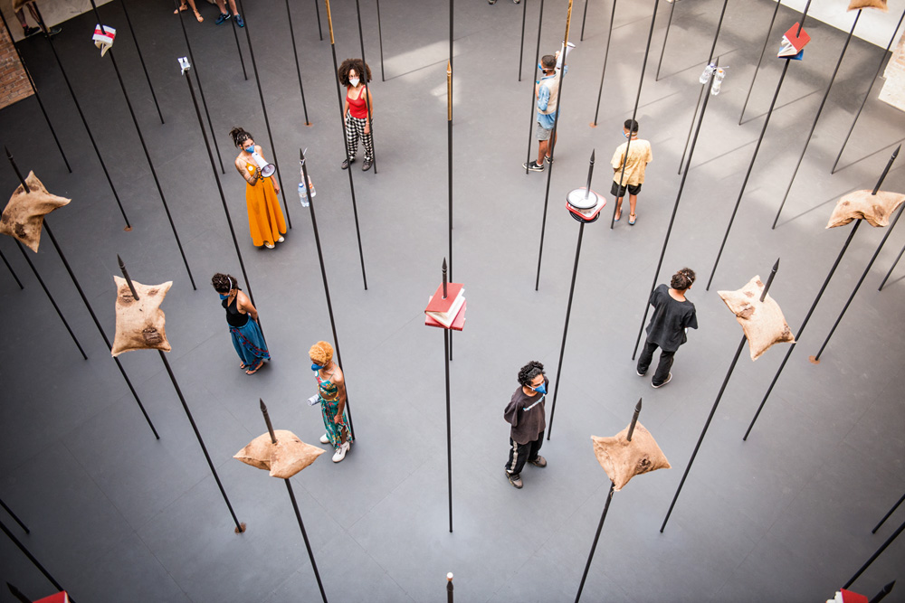 instalação noite longa fotografada de cima, em que itens do cotidiano são fincados nas pontas das lanças; membros de um coletivo estão um de costas para o outro, em formato de circulo, dentro da instalação, durante performance de crítica social