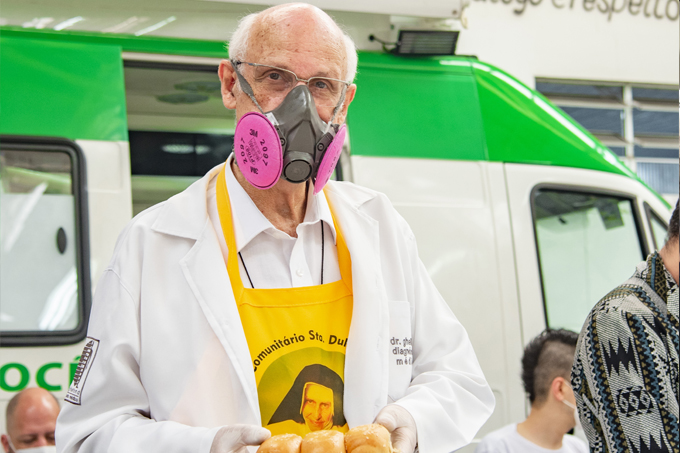 Padre Júlio Lancelotti de máscara, avental amarelo sobre jaleco branco distribuindo pães em galpão.