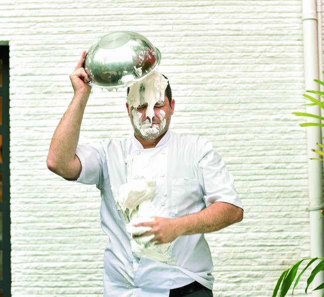 O confeiteiro Rafael Protti em frente à parede branca de dólmã segurando tigela de metal com claras em neve caindo sobre a sua cabeça.