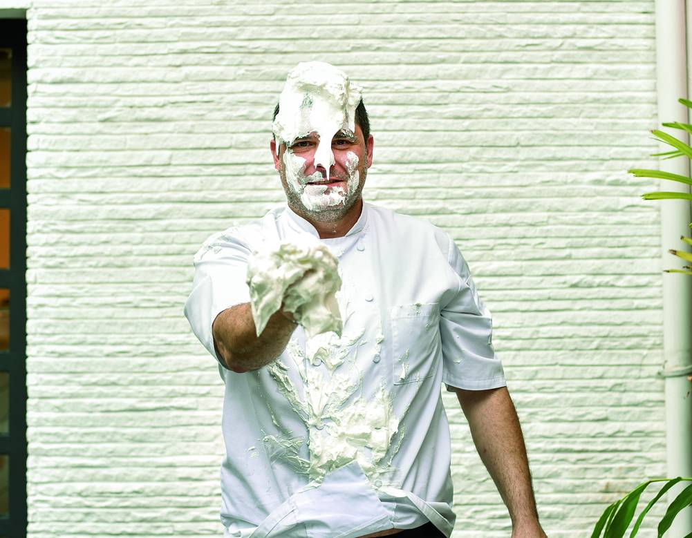 COMER & BEBER 2021 – Rafael Protti O confeiteiro Rafael Protti em frente à parede branca de dólmã todo sujo de claras em neve segurando uma parte oferecendo para a foto.