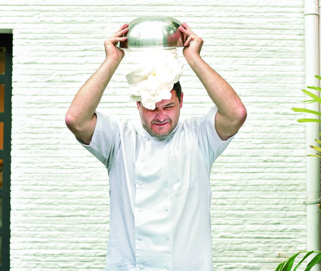 Rafael Protti = Crime Pastry Shop O confeiteiro Rafael Protti em frente à parede branca de dólmã segurando tigela de metal com claras em neve caindo sobre a sua cabeça.