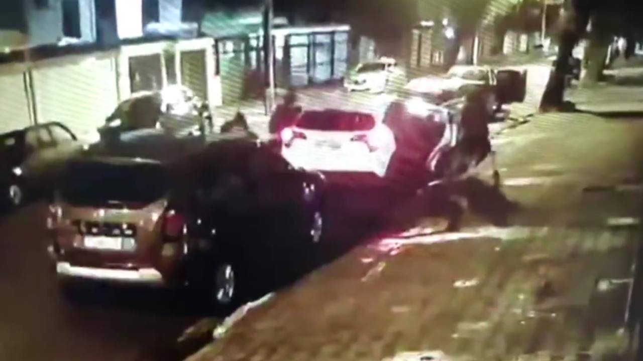 A imagem mostra um carro parado e outros quatro homens encapuzados em volta dele