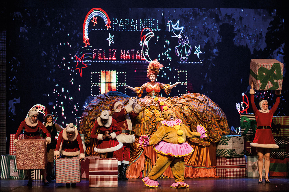 Em um palco de teatro, há pessoas de Papai Noel, decorações de Natal, presentes, luzes...