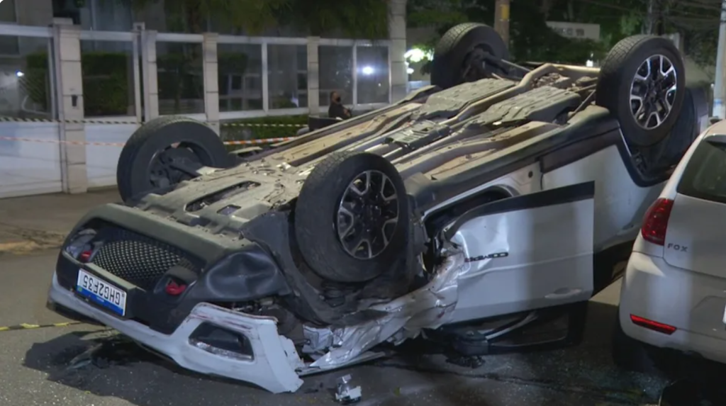 Imagem mostra carro branco capotado, com as rodas para cima no meio de uma rua.