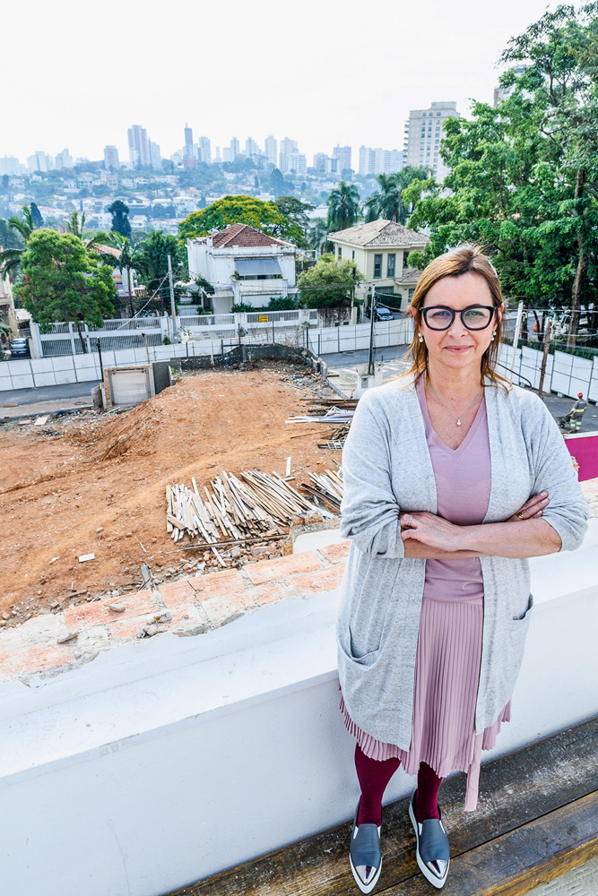 Carla Pernambuco (LUCAS TERRIBILI – DIVULGAÇÃO) Carla Pernambuco posa de braços cruzados com cardigã cinza e vestido rosa. Ao fundo, espaço com cor de terra destaca obras do metrô.