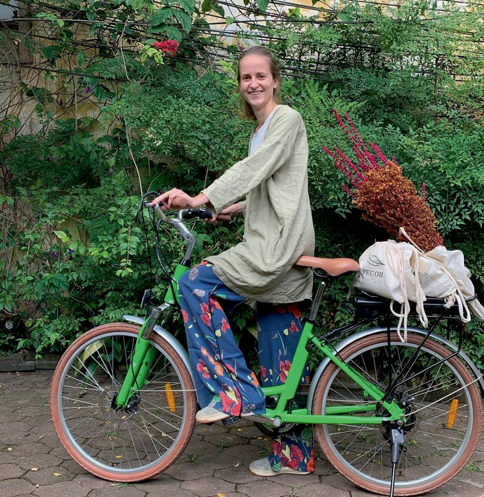 Elise-PECOH-Entregas-de-Bicicleta.jpg (1) A imagem mostra Elise em cima de uma bicicleta, apoiada com um pé. Ela sorri para a câmera em um ambiente cheio de plantas.