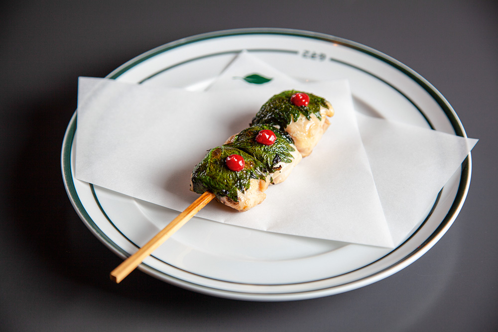 Kotori Espetinho com três pedaços de frango sobre prato de louça branca coberto por pesto e gota de molho vermelho.