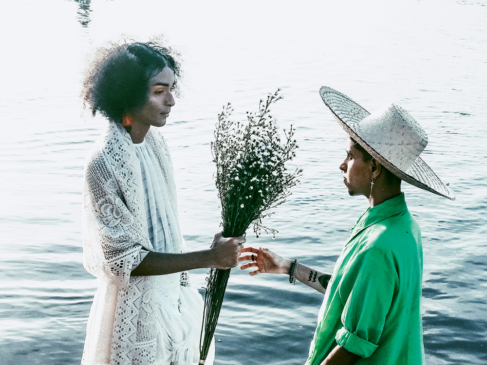 IMG_0890.JPG Imagem mostra duas pessoas, uma de vestido branco e outra de cmaisa verde, se olhando enquanto uma entrega flores à outra. No fundo, o mar.