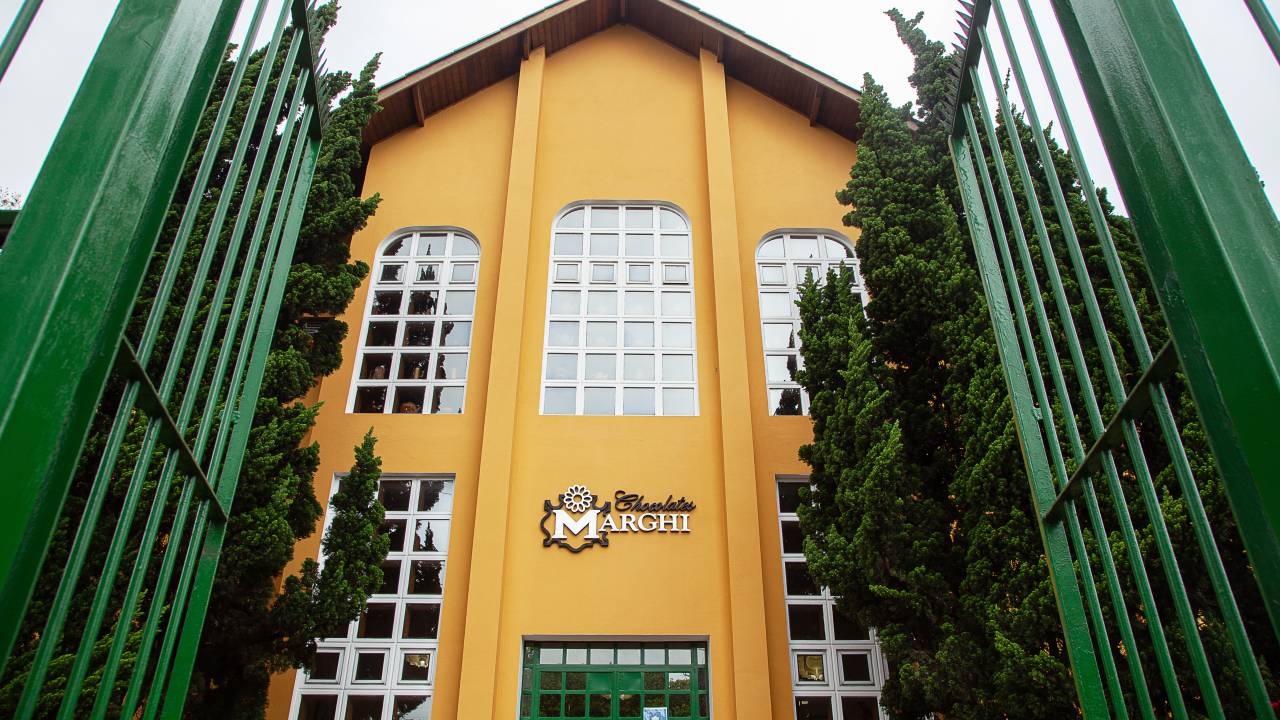 Fachada amarelo-mostarda da Chocolates Marghi ao lado de árvores verdes.