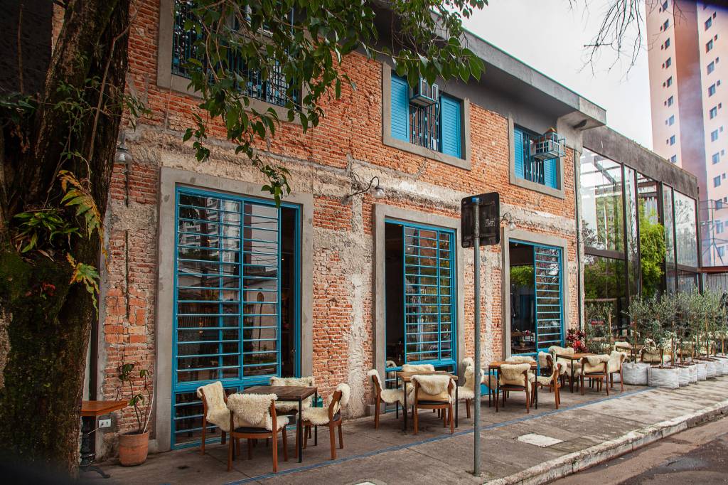 Achados ELO – Quintana Fachada do bar Quintana com paredes de tijolinhos e mesas na calçada com cadeiras cobertas por cobertores de pelo.