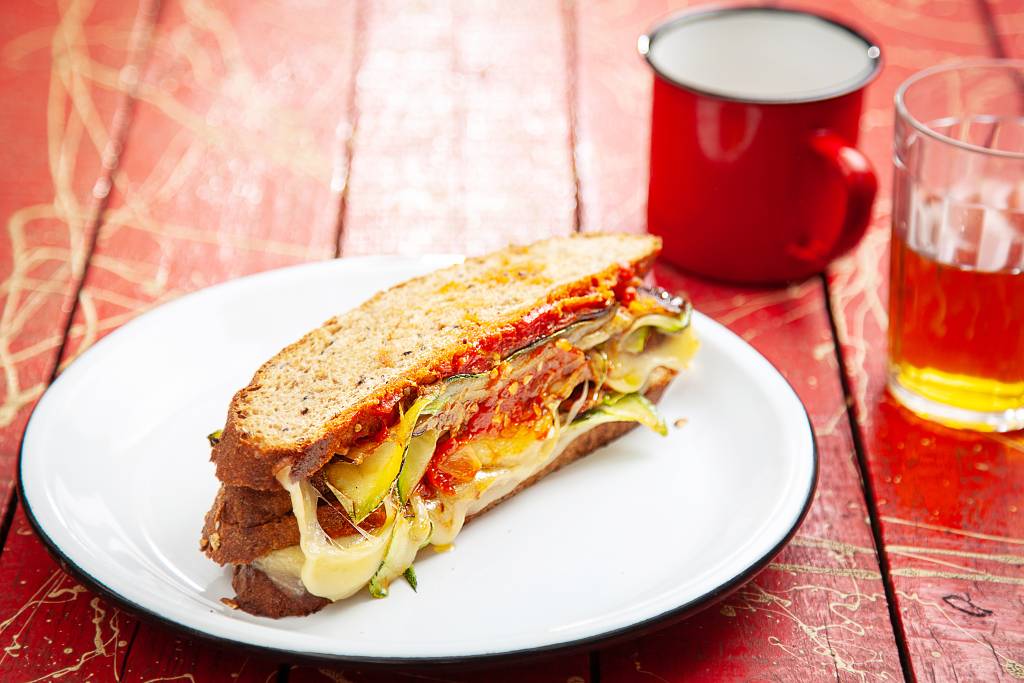 Achados Elo – Miolo Padaria Artesanal Sanduíche com queijo e molho de tomate sobre prato branco em cima de mesa de madeira pintada de vermelho.