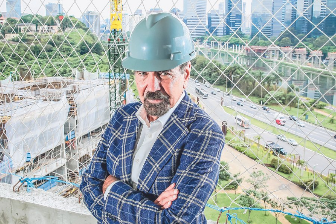 Homem de braços cruzados, vestido com camisa xadrez azul e capacete de proteção cinza, com uma construção e a Marginal Pinheiros atrás.