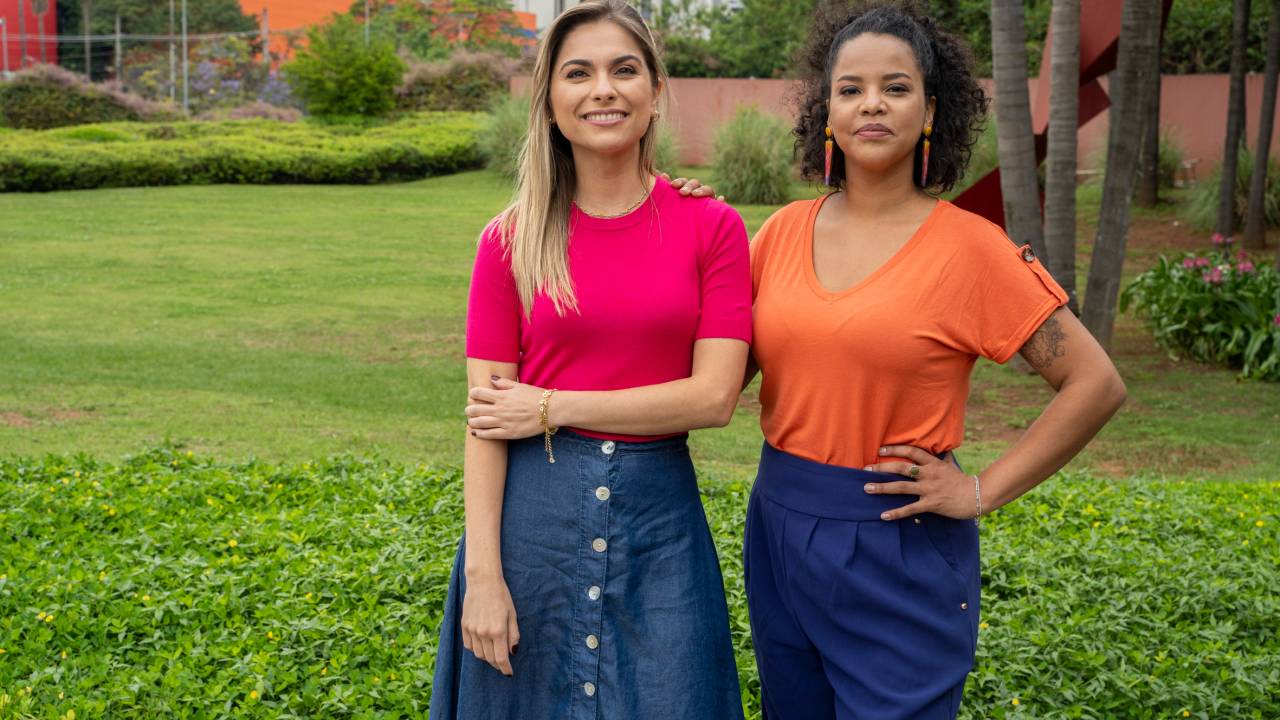 As apresentadoras do programa Mistura Paulista: Luiza Vaz e Denise Thomaz Bastos, que posam lado a lado vestindo jeans e camisetas nas cores rosa e laranja. Ao fundo, um grande jardim.
