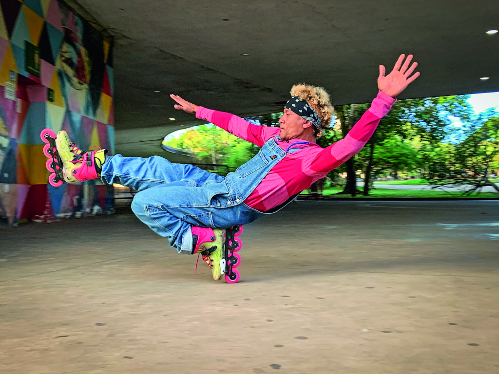 Homem de patins, macacão jeans e camiseta listrada vermelha e rosa apoiado com um pé só e em posição horizontal.