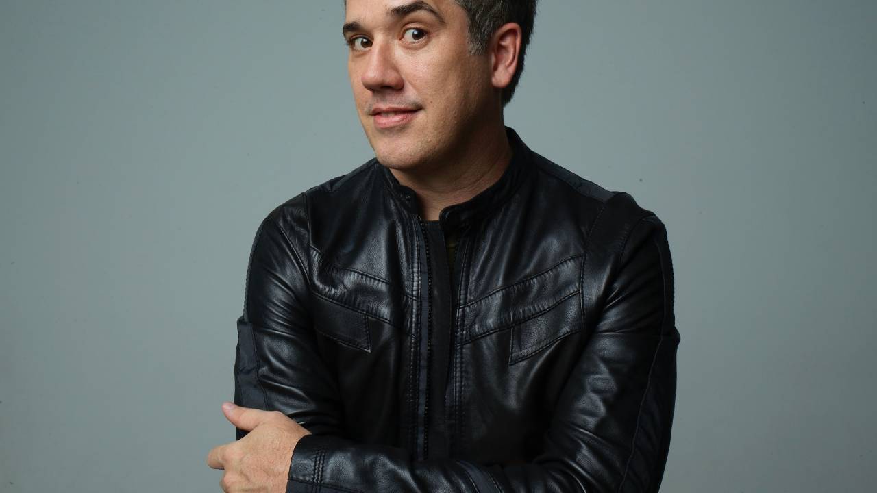 Rogério Flausino, vocalista do Jota Quest, posa de jaqueta de couro, sobrancelha erguida e braços cruzados.