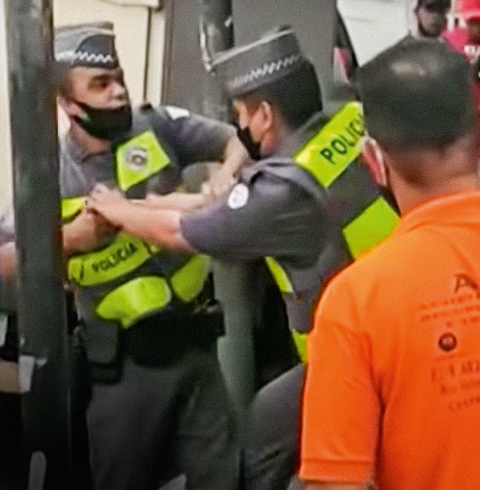Policial aponta arma para outro A foto mostra um PM agindo contra o outro, na tentativa de desarmar o que estava apontando uma arma para a sua cabeça. Há pessoas olhando