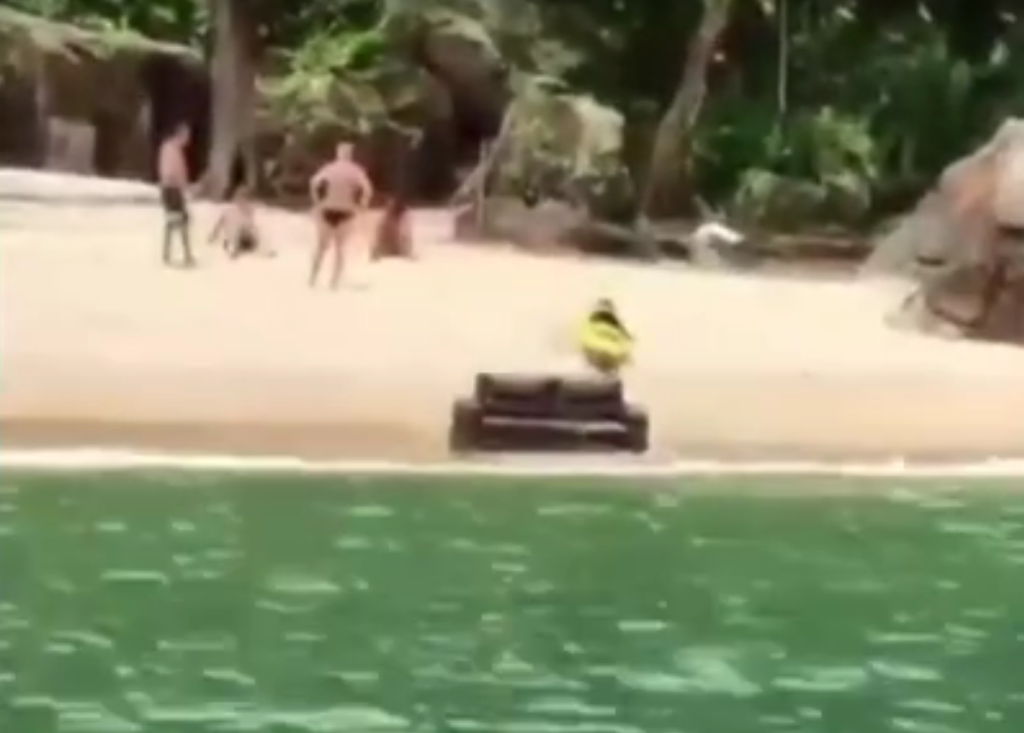 A imagem mostra um sofá na beira de uma praia, próximo a água do mar. Em volta, há banhistas observando o objeto.