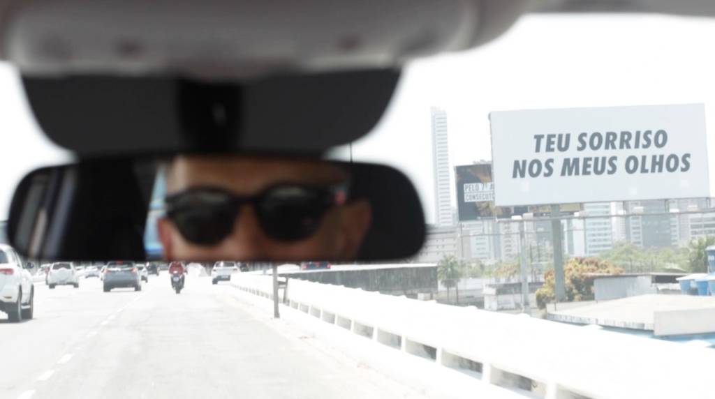 Foto exibe retrovisor com Felipe usando óculos escuros. Ao fundo, outdoor com a frase "teu sorriso nos meus olhos".