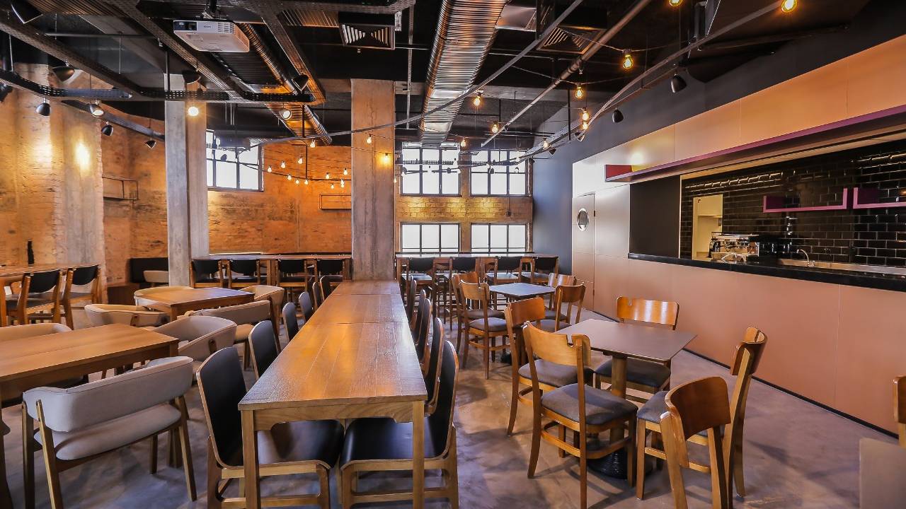 O salão do Boteco do 28 com mesas e cadeiras de madeira e janela para a cozinha à direita.