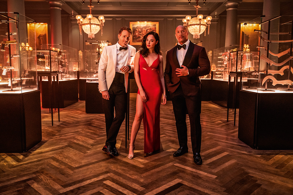 A imagem mostra Gal Gadot, Ryan Reynolds e Dwayne Johnson em um salão chique, usando roupas de gala.