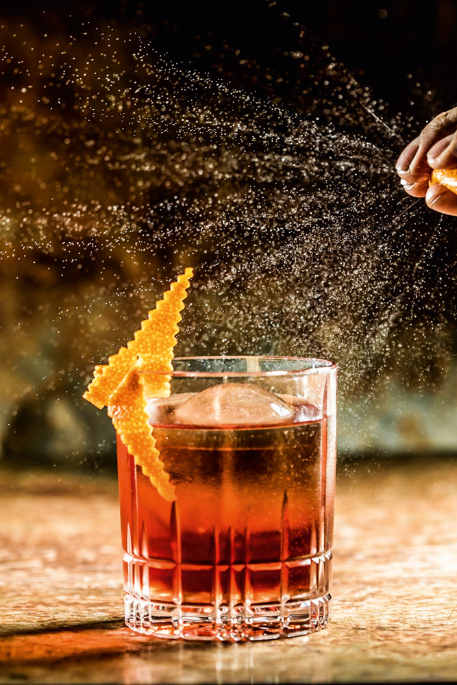 Balaio IMS Drinque de cor alaranjada servido em um copo de fundo redondo e decorado com uma parte da casca de uma laranja
