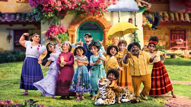 encanto – credio Disney.jpg Imagem mostra animação de uma grande família vestindo roupas brancas, vermelhas, azuis e amarelas, posando para a foto juntos na frente de uma vila repleta de flores.