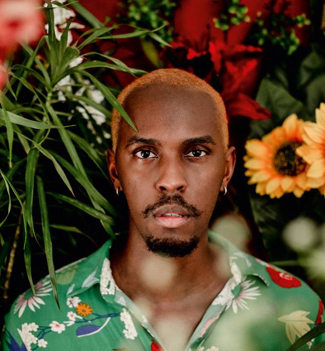 Retrato de Stefano Volp. Ele é um homem negro, de barba escura e cabelo crespo platinado. Ele usa uma camiseta estampada de botões e um brinco em cada orelha. No pano de fundo é possível ver plantas e um girassol