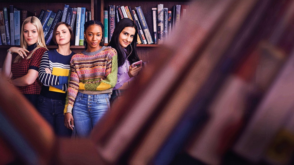 Imagem mostra quatro meninas posando e sorrindo para a câmera em uma biblioteca. Ao fundo, uma estande de livros.