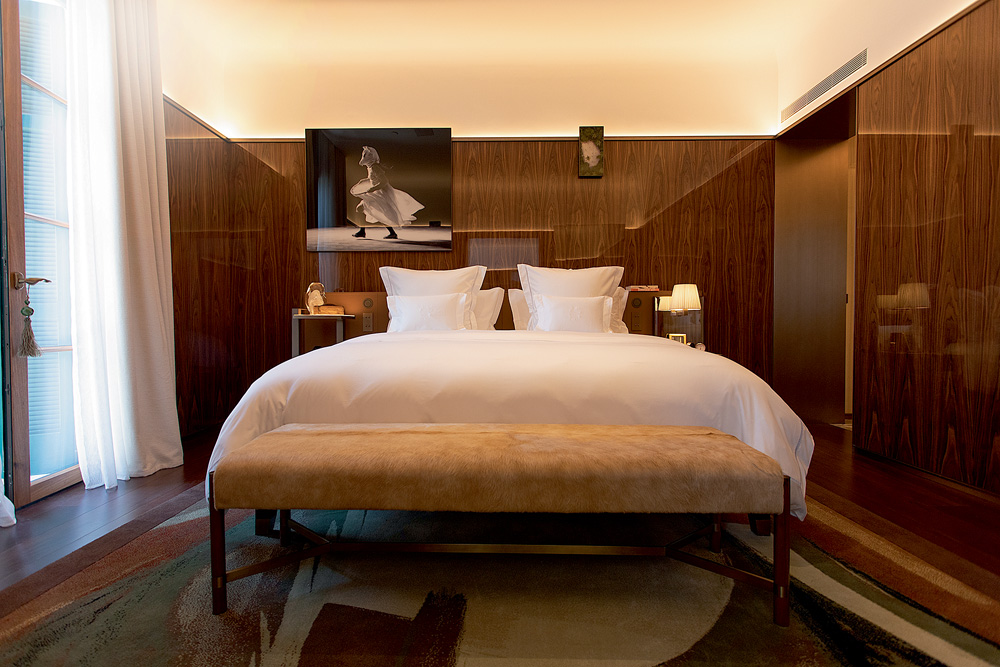 CIDADE MATARAZZO Foto mostra um quarto de hotel. Uma cama branca é o centro da foto. Paredes escuras e com obras penduradas