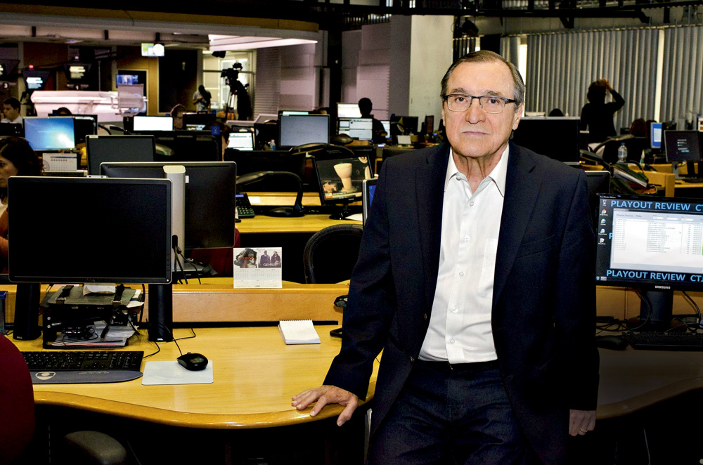 O âncora Carlos Alberto Sardenberg posa de terno em uma redação com vários computadores