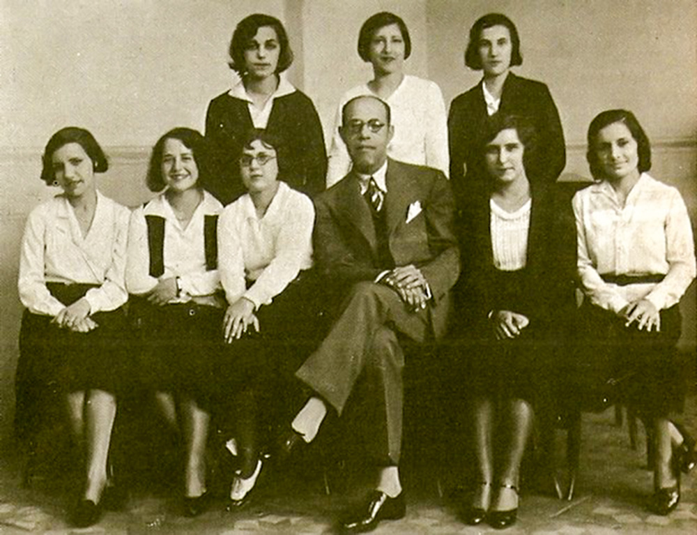 Crédito_Domínio Público.jpg Imagem em branco e preto mostra 8 mulheres sentadas e em pé em volta de um homem, ao centro, de terno e sentado de pernas cruzadas.