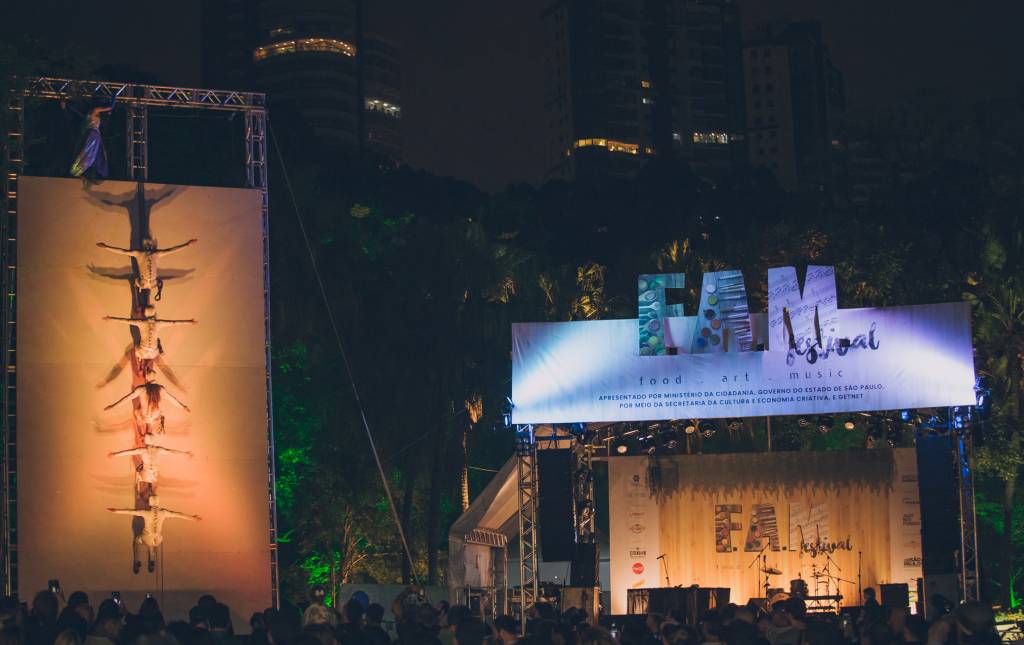 FAM0727184048 (1) Imagem mostra um painel à esquerda com várias pessoas de roupas brancas penduradas, uma embaixo da outra. À direita, um palco iluminado. A foto foi tirada de noite.