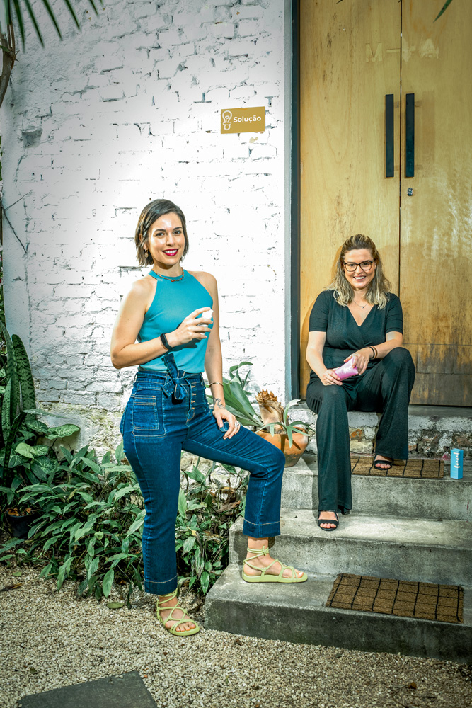 Imagem mostra duas mulheres, uma em pé, com um dos pés em uma escada. A outra, sentada na escada.