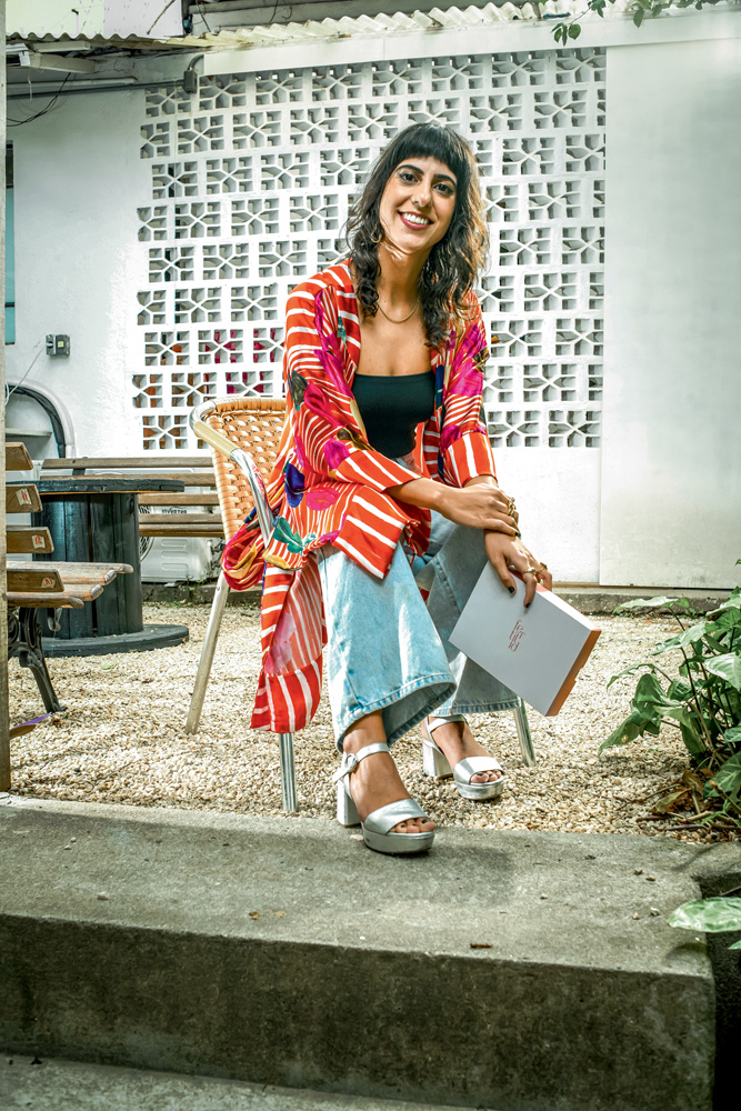São Paulo 03 dezembro 2021FEMTECHS Foto Alexandre Battibugli VSP Imagem mostra mulher sentada vestindo roupas coloridas. Ela sorri para a câmera e segura uma caixa branca com uma das mãos.