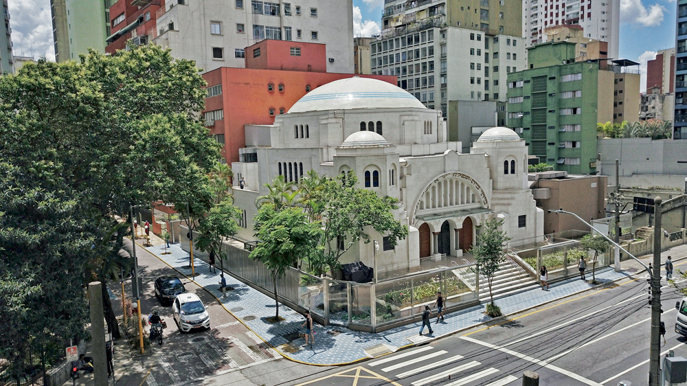 Uma foto um pouco longe de uma antiga sinagoga, com algumas árvores em volta e em um cruzamento