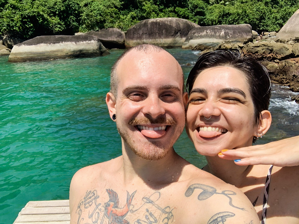 Léo e Leticia: amor na pandemia Um casal posa para a selfie em frente a um mar esverdeado em Paraty