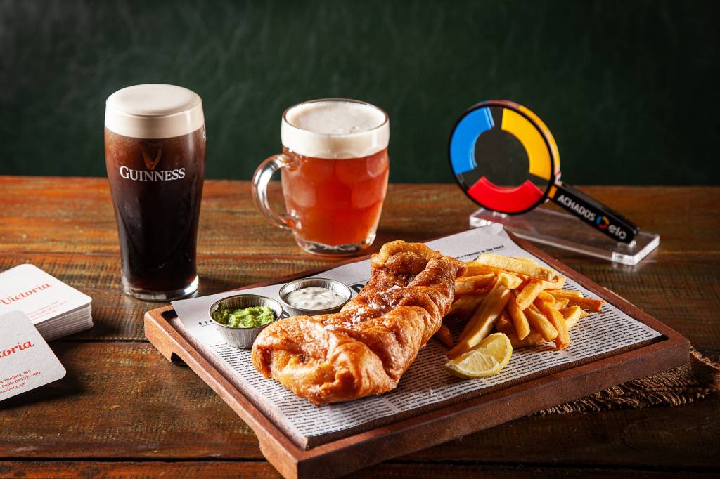 Achados ELO – The Victoria Tábua com fish & chips sobre mesa de madeira ao lado de copo e caneca de chope.