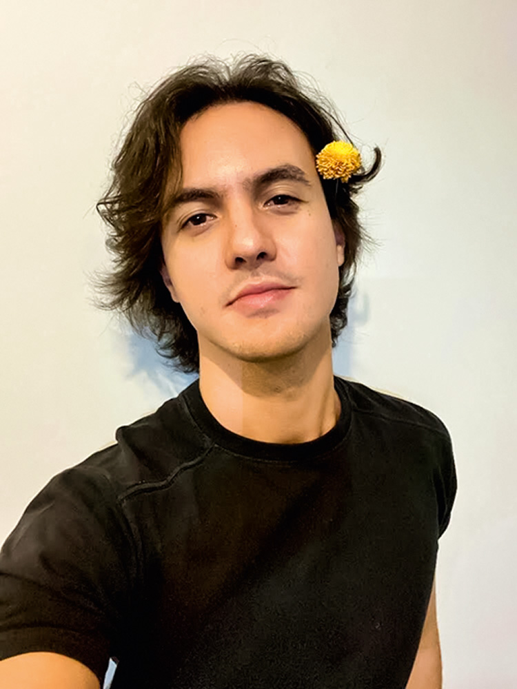 IMG_4186.jpeg Foto de Renato. Ele é um homem branco, sem barba, de cabelos compridos e escuros. usa uma camiseta preta e uma flor amarela no canto da orelha.
