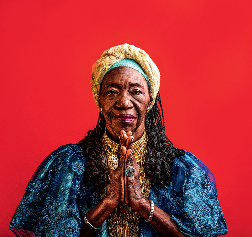 Lia de Itamaraca_foto_por_Jose_de_Holanda_ Imagem mostra mulher com roupa azul e traços dourados sobre fundo vermelho. Ela faz está com as duas palmas levantadas e se encostando.