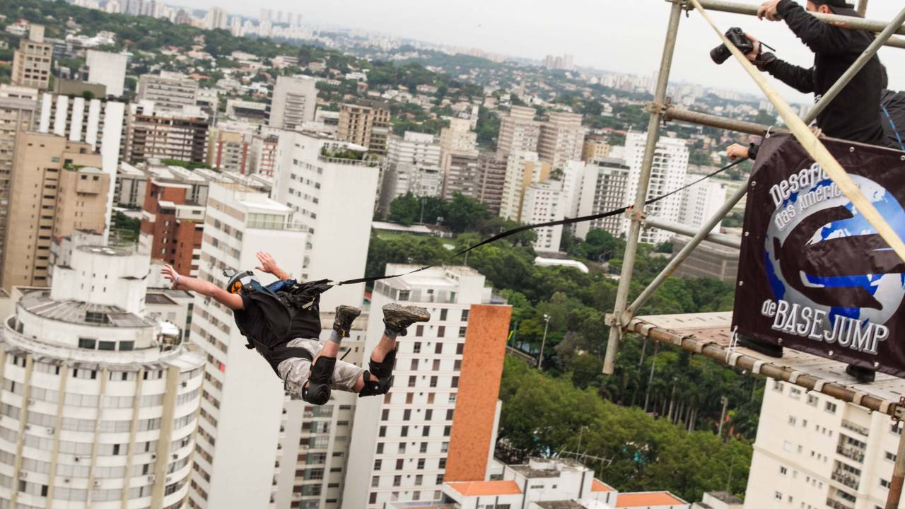 Imagem mostra homem saltando de paraquedas. Abaixo, via movimentada do centro de São Paulo