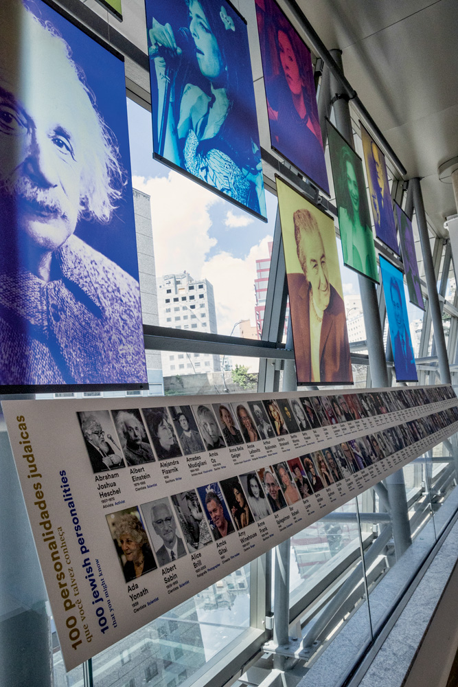 Várias fotos coloridas de personalidades judias estão apoiadas em janelas. Há um painel com 100 pessoas judias também perto das janelas