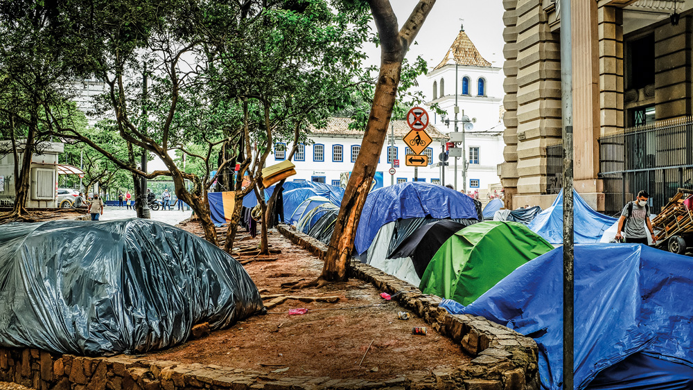 Imagem mostra diversas barracas azuis e verdes ao lado de um canteiro. Ao fundo, um prédio antigo.